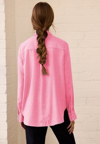 Woman with long braided hair wearing a loose, pink and white striped shirt and dark pants, standing against a pale brick wall.