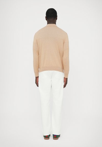 Világos bézs kötött pulóver alacsony nyakkal, hosszú ujjakkal és szoros szabással. Fehér nadrággal és barna cipővel párosítva, zöld részletekkel.