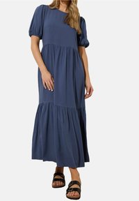 Robe maxi fluide bleue avec des manches bouffantes, un col rond et une jupe à volants. Portée avec des sandales noires à lanières. Tissu doux avec une coupe décontractée.