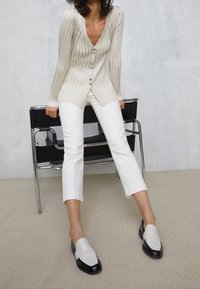 Crème ribgebreide cardigan met een V-hals en knoopsluiting, gedragen over witte cropped broeken. Zwart-beige instappers zorgen voor contrast. Minimalistisch ontwerp.