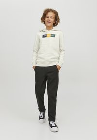 Felpa grigia chiara con logo, tasca a marsupio frontale e cappuccio con cordino; abbinata a pantaloni neri e sneakers nere.