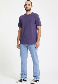 T-shirt di cotone viola a maniche corte, abbinata a jeans di denim azzurro chiaro e sneaker bianche e nere; in piedi su uno sfondo neutro.