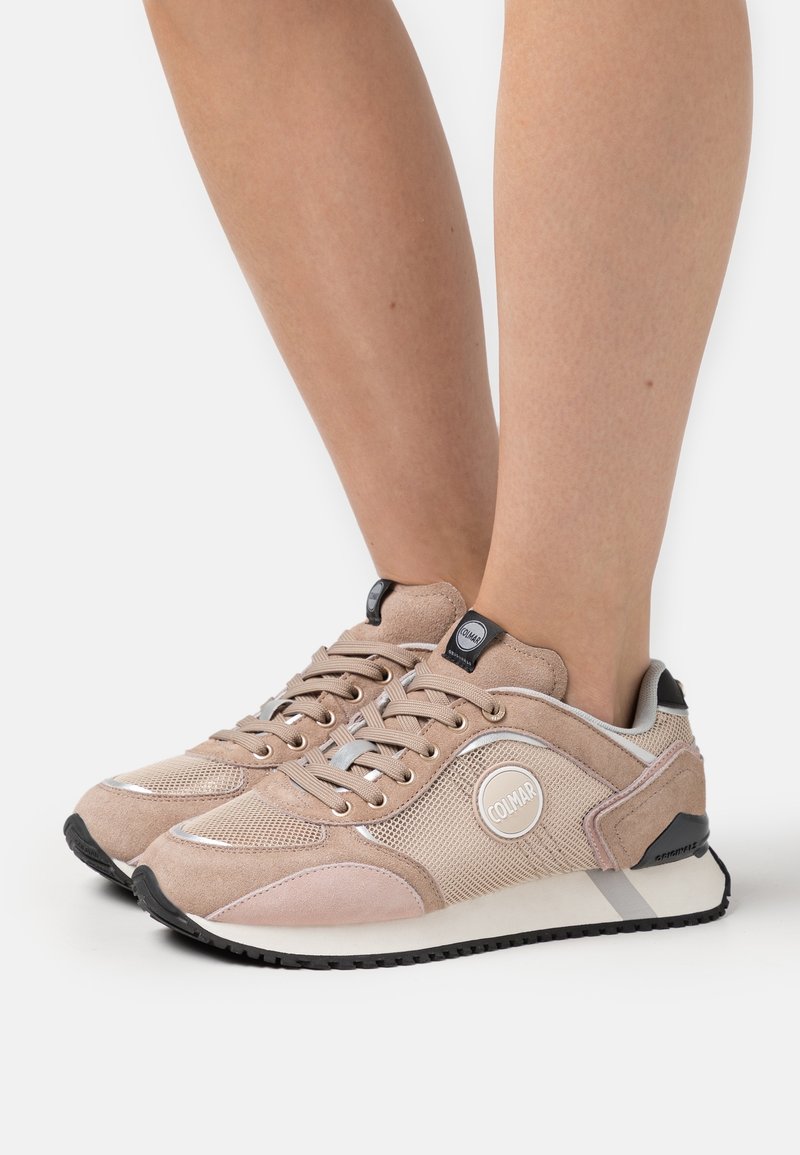 Béžové semišové a síťované tenisky s černou podrážkou, stříbrnými akcenty a kulatým logem na boku. Šněrovací design a polstrovaný límec.