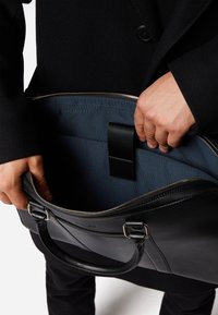Sort lædertaske med lynlås. Indvendigt er tasken beklædt med blåt stof og har et centralt bånd. Den har to håndtag og detaljer i sølvfarvet hardware.