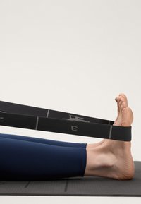 Čierny pretiahnutý popruh je položený okolo nahých nôh, ležiacich na šedej jogovej podložke. Nohy sú pokryté modrými športovými legínami, ktoré ukazujú hladké textúry.