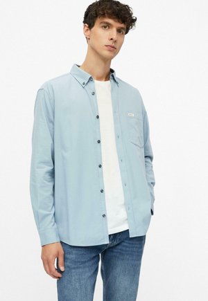 Junger Mann trägt hellblaues Hemd über weißem T-Shirt und blauen Jeans, steht mit einer Hand in der Tasche vor weißem Hintergrund.