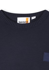 Granatowy sweatshirt z prążkowanym okrągłym dekoltem, metką z tkaniny oraz małą gumową łatką Timberland na lewej piersi. Gładka tekstura.