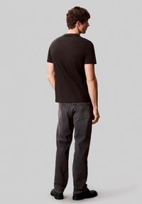 Camiseta de algodón negra con un corte clásico, combinada con jeans gris oscuro que presentan detalles en los bolsillos y costuras mínimas, usada con zapatos negros.