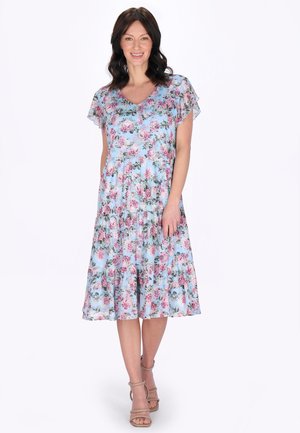 Vrouw in een knielange, lichtblauwe bloemenjurk met zwierige mouwen, staand en glimlachend tegen een witte achtergrond.