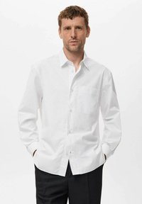 Camisa blanca de manga larga, con botones, con rayas verticales, un cuello puntiagudo y un bolsillo en el pecho. Textura suave, corte ajustado y tejido ligero.