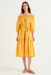 Gul off-shoulder klänning med volangkrage, ballongärmar, bältad midja och lager-på-lager kjol; bomullstyg, slät textur.