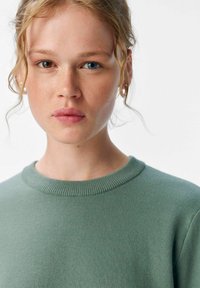 Jeune femme aux cheveux blonds bouclés et à l'hétérochromie, portant un pull vert, regardant directement l'objectif sur un fond blanc.