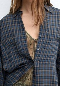 Chemise à carreaux bleue et brune avec une texture douce et une fermeture à boutons, portée par-dessus un vêtement transparent beige avec un détail de nouage au niveau du col.