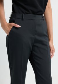 Femme portant un pantalon noir sur mesure et un haut noir, avec la main gauche dans la poche et la main droite détendue sur le côté.