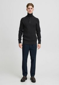 Pulover negru din tricot cu un guler înalt, cu trei butoane, asortat cu pantaloni închiși la culoare și mocasini negri, stând în fața unui fundal neutru.