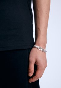 Camiseta de algodón negra con una etiqueta lateral, combinada con una pulsera de cadena plateada en una mano. Diseño simple, textura suave, forma ajustada.