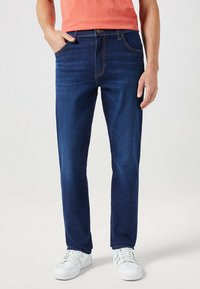 Vaqueros azul oscuros de denim, con un corte recto, diseño de cinco bolsillos y costuras visibles. Llevados con zapatillas blancas.