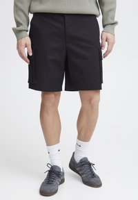 Schwarze Cargo-Shorts mit Seitentaschen, aus einem strapazierfähigen Stoff gefertigt. Getragen mit grauen Sneakers und weißen Knöchelsocken, vor einem weißen Hintergrund präsentiert.