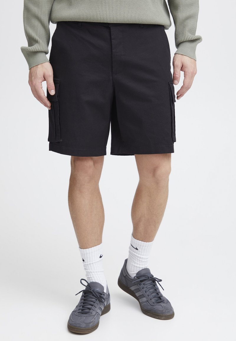 Schwarze Cargo-Shorts mit Seitentaschen, aus einem strapazierfähigen Stoff gefertigt. Getragen mit grauen Sneakers und weißen Knöchelsocken, vor einem weißen Hintergrund präsentiert.