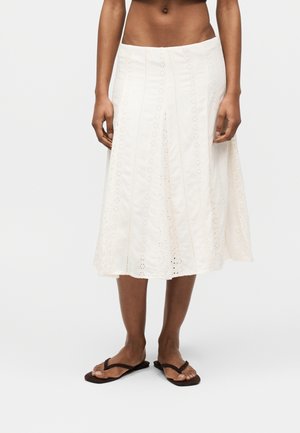 Weißer Midirock mit Augelloch-Blumenstickerei, getragen mit schwarzen Flip-Flops an einer dunkelhäutigen Person vor einfachem Hintergrund.