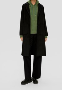 Zwarte wollen overjas met voorkantenzakken, gedragen over een groene hoodie. Model draagt zwarte wijde broeken en stevige zwarte laarzen.