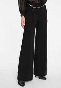 Zwarte brede broek gemaakt van zacht materiaal met een gladde textuur. Voorzien van een metallic riem met kettingdetail. Gecombineerd met zwarte hakken.