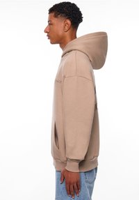 Sweatshirt beige à capuche avec une poche kangourou, des poignets côtelés et un logo discret sur la poitrine. Matière douce et coupe décontractée.