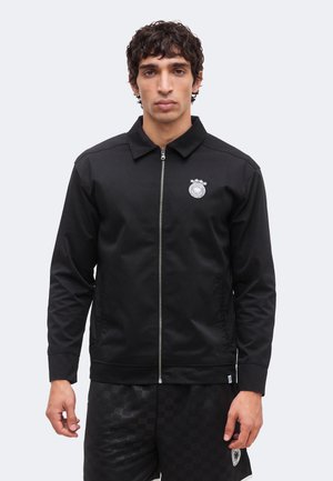 Nuori mies kiharat hiukset, musta vetoketjutakki, jossa pieni rintamerkki, ja mustat ruudulliset shortsit.