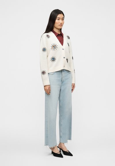 Κρεμ ζακέτα με μαύρη και έγχρωμη κεντητική σε σχέδιο ηλιαχτίδας, συνδυασμένη με ανοιχτό μπλε τζιν με φαρδύ πόδι και μαύρα μυτερά παπούτσια.