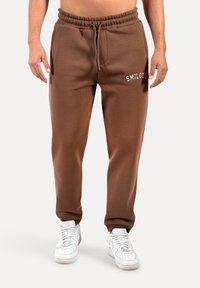 Brune sweatpants med elastisk talje og snor, med en frontlomme og hvid broderet tekst "SMILODO," parret med hvide sneakers.