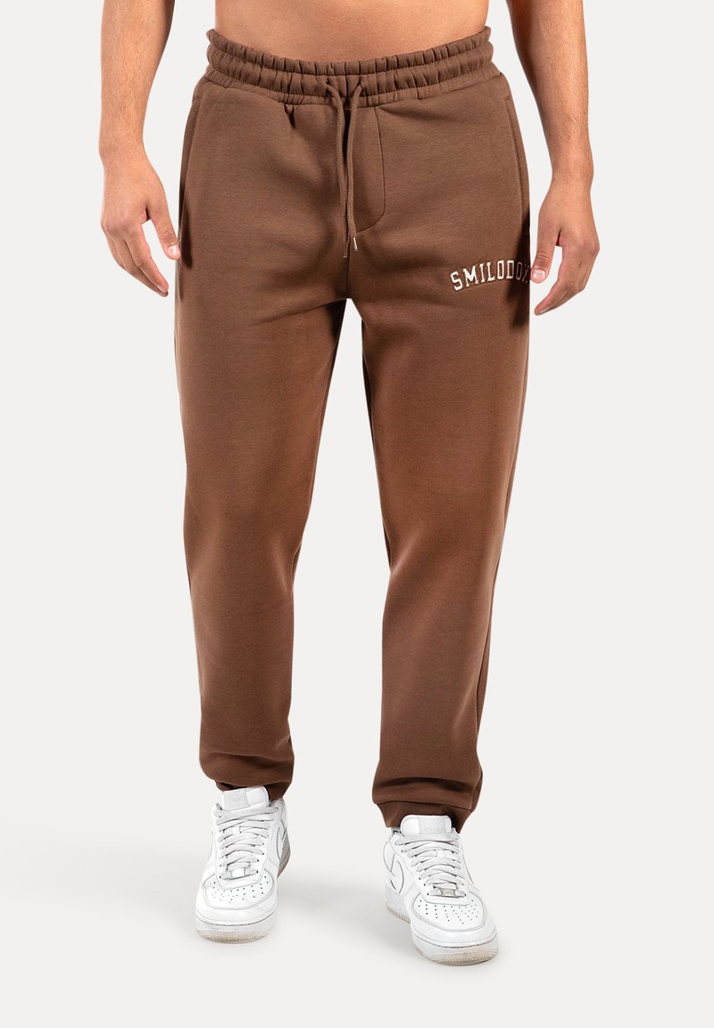 Brune sweatpants med elastisk talje og snor, med en frontlomme og hvid broderet tekst "SMILODO," parret med hvide sneakers.