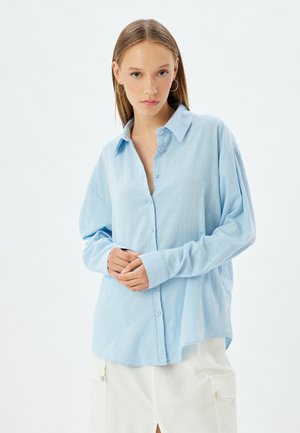 Chemise bleu clair à boutons en tissu doux et texturé, avec des manches longues et une coupe décontractée, agrémentée de boutons bleu clair.