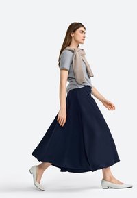 Haut gris à manches courtes associé à une longue jupe navy, complété par un wrap en tricot beige. Modèle portant des ballerines crème. Fond uni.