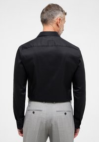Maglietta nera a maniche lunghe con una superficie liscia, dotata di colletto classico e polsini abbottonati, abbinata a pantaloni grigi chiari su misura con tasche.