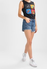 Camiseta sin mangas negra con paneles gráficos coloridos de temática espacial, combinada con pantalones cortos de mezclilla con dobladillos deshilachados y zapatillas altas blancas.