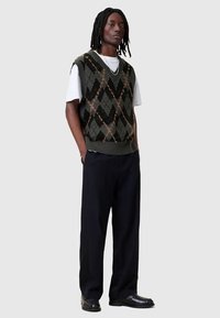 Colete de malha em verde escuro com padrão argyle em tons de preto e laranja, usado por cima de uma t-shirt branca, combinado com calças e sapatos pretos.