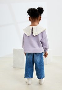 Felpa lilla con un grande colletto bianco rifinito in pizzo, abbinata a jeans larghi blu e scarpe da ginnastica bianche. Bambino visto da dietro.