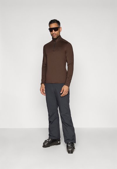 Brun långärmad zip-halströja som kombineras med mörkgrå skidbyxor och svarta skidskor. Slät textur och åtsittande design.