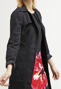 Černý semišový dlouhý kabát s límcem, který má volný střih. Nalezeno přes živě květinově vzorované šaty v červených a růžových odstínech.