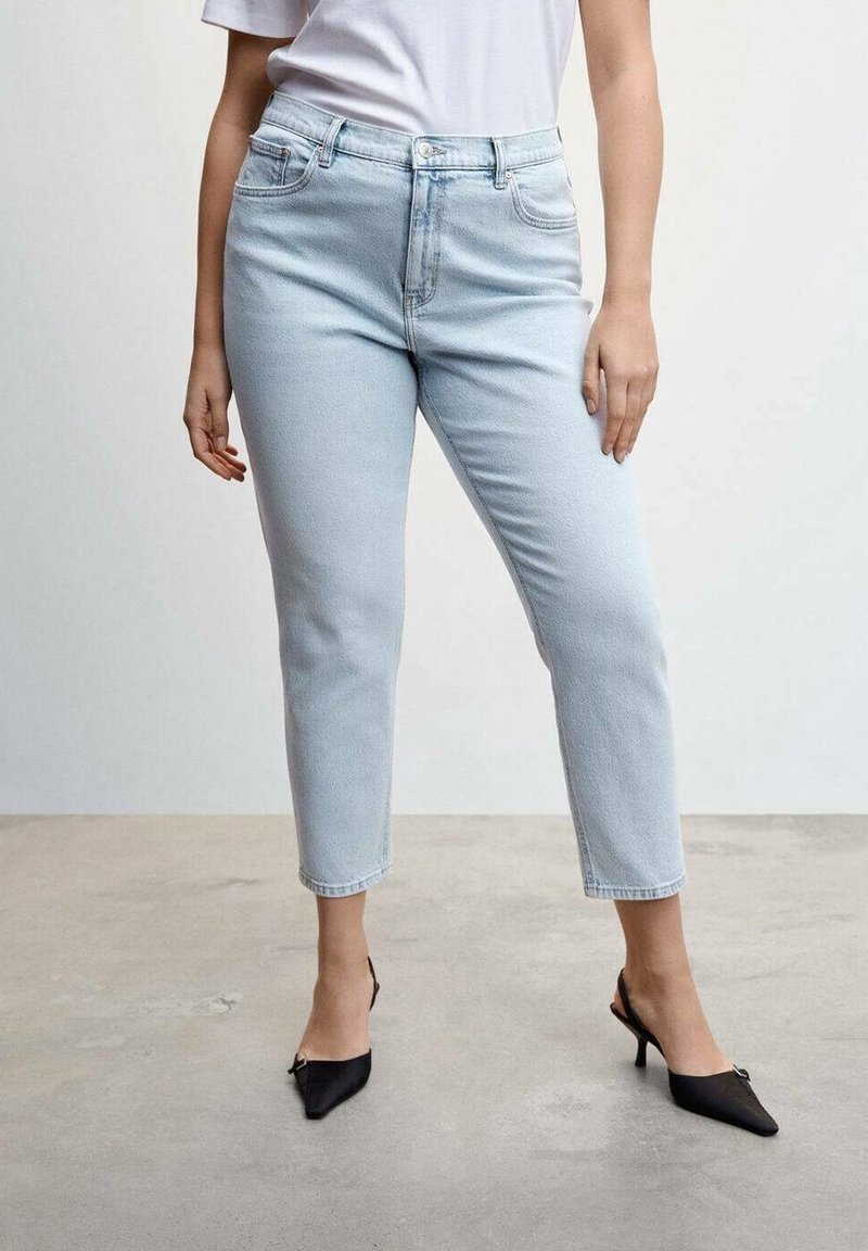 Femme portant un jean bleu clair taille haute et des talons noirs pointus, debout sur un sol en béton simple contre un mur blanc.
