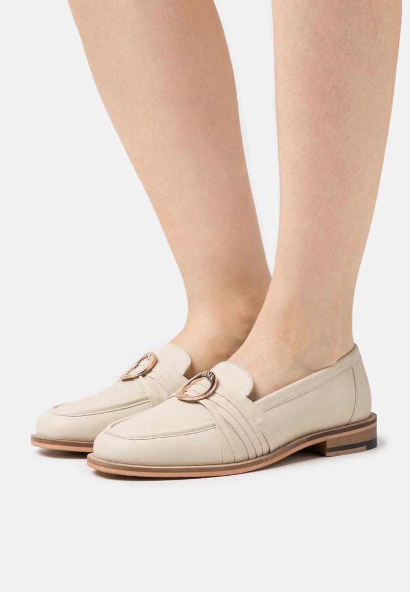 Mocassins en cuir beige avec un bout rond, des coutures décoratives et un accent métallique sur la bride. Talon bas, texture lisse.