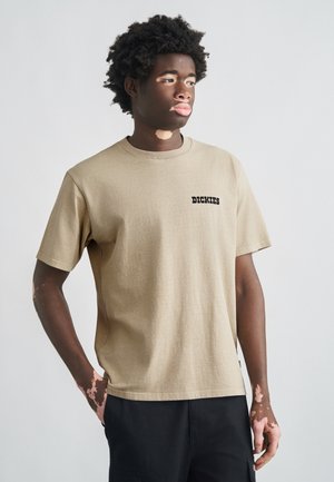 Μπεζ βαμβακερό μπλουζάκι με κοντά μανίκια και στρογγυλή λαιμόκοψη. Η λέξη "DICKIES" είναι τυπωμένη με μαύρα γράμματα στο αριστερό στήθος. Ομοιόμορφη υφή, άνετη εφαρμογή.