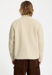 Veste en polaire couleur crème avec un col montant. Tissu doux et texturé. Le dos n'a pas de fermetures visibles, portée avec un pantalon en corduroy marron.