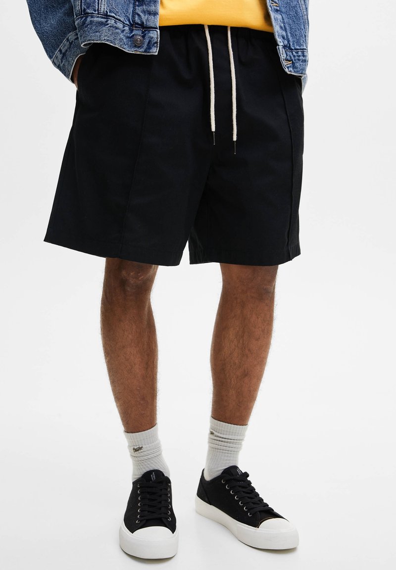 Homme portant un short noir à cordon, des chaussettes blanches, des baskets noires, un t-shirt jaune et une veste en jean, les mains dans les poches.