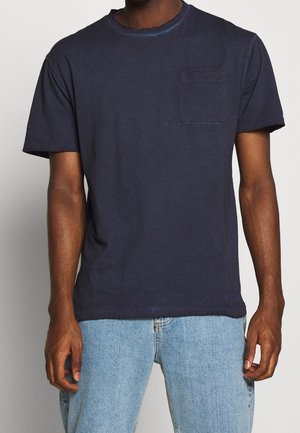 Mann trägt ein schlichtes dunkelblaues T-Shirt mit kurzen Ärmeln und Brusttasche sowie hellblaue Jeans, steht vor einem schlichten Hintergrund.