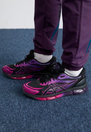 Baskets de sport noires avec des accents dégradés rose et violet, tige en mesh, semelle épaisse et lacets noirs, accompagnées de chaussettes blanches et de pantalons violets.