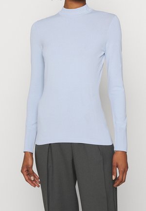 Femme portant un pull à col roulé ajusté bleu clair à manches longues et un pantalon plissé gris foncé, debout devant un fond uni.
