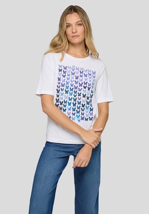 Frau trägt weißes T-Shirt mit mehrfarbigem blau-lila Schmetterlingsdruck, kombiniert mit blauen Jeans, steht vor grauem Hintergrund.