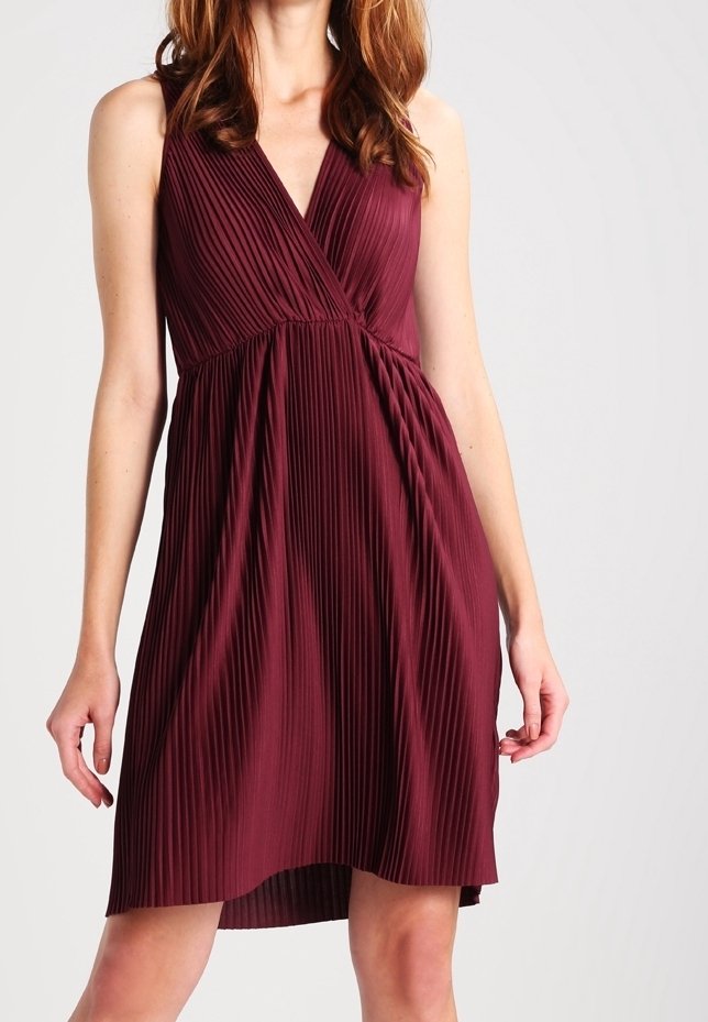 Femme portant une robe bordeaux sans manches, plissée, avec un col en V et une taille froncée sur un fond neutre.