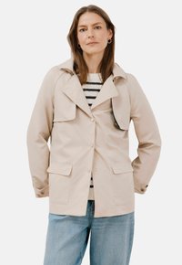 Beige, knäppt jacka med krage, två framfickor och ett lager av bröstfickan. Bärs över en randig tröja och ljusblå jeans.